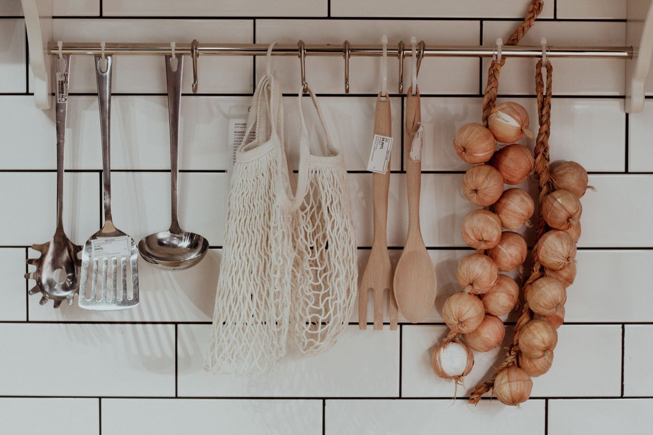 Rustic kitchen setup with utensils and braided onions on wall hooks against tiled background.